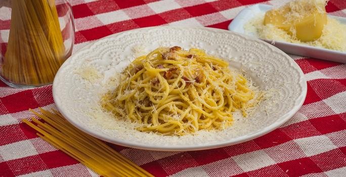 Foto de uma mesa com uma toalha xadrez vermelha e branca. Sobre ela há um prato branco com a receita de Spaghetti à Carbonara, alguns macarrões crus espalhados e num pote e, do lado direito, um pedaço de queijo com queijo ralado.