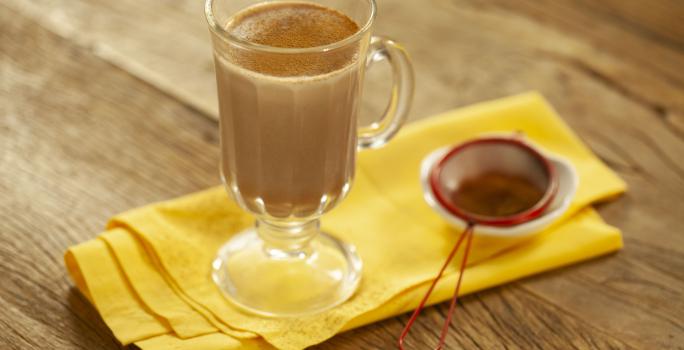 Fotografia em tons de marrom e amarelo de uma bancada de madeira com um paninho amarelo, sobre ele uma xícara transparente com leite, chocolate baton derretido e canela em pó. Ao lado uma peneira vermelha com canela.