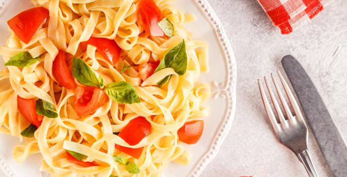 fotografia em tons de branco, amarelo e vermelho de uma bancada branca vista de cima. Contém um prato redondo branco com macarrão tipo talharim com pedaços de tomate e folhas de manjericão. Ao lado garfo e faca para servir.