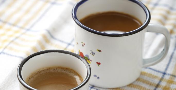 Fotografia em tons de branco, bege e azul, com duas canecas brancas com borda azul contendo bebida em tons café-com-leite, sobre guardanapo branco com listras azul e bege, tudo sobre bancada de madeira.