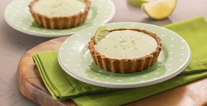 Foto da receita de tortinha de limão. Observa-se dois pratos verdes de sobremesa com as tortinhas sobre um guardanapo verde e uma tábua de madeira. Atrás, duas fatias de limão.