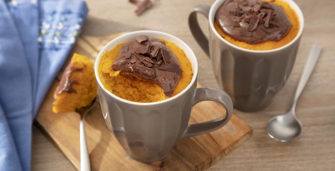 Fotografia em tons de laranja em uma bancada de madeira clara, uma tábua de madeira, um paninho azul, duas canecas marrons com o bolo de cenoura dentro de cada uma e por cima o brigadeiro de Nescau.