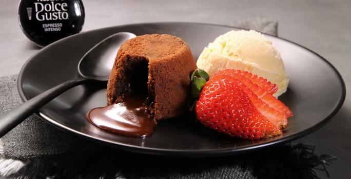 Fotografia em tons de preto em uma bancada de madeira escura, um prato preto redondo com o petit gateau de café em cima, ao lado, uma bola de sorvete de creme e um morango para decorar. Ao fundo, uma cápsula do café intenso de Dolce Gusto.