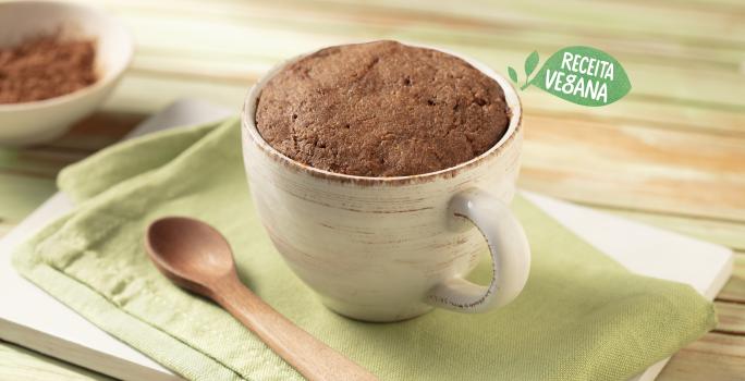 Fotografia em tons de verde, branco e marrom de uma bancada de madeira clara com uma tábua branca e um paninho verde, sobre ele uma caneca branca com o bolo de cacau e uma colher de madeira. Ao fundo um recipiente pequeno redondo branco com cacau em pó.