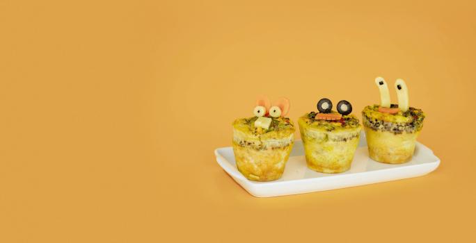 Fotografia em tons de amarelo em uma mesa e fundo amarelo escuro, um recipiente branco retangular e três mini muffins de omelete com desenhos infantis.