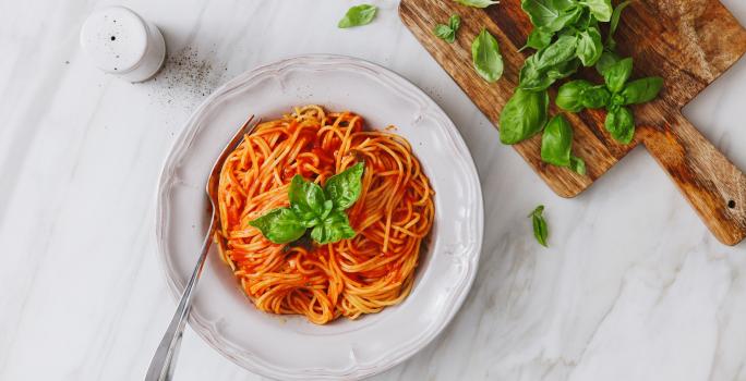 fotografia em tons de branco e vermelho de uma bancada branca vista de cima. Ao centro um prato branco com macarrão espaguete e molho de tomate, e ao lado uma tábua com folhas.