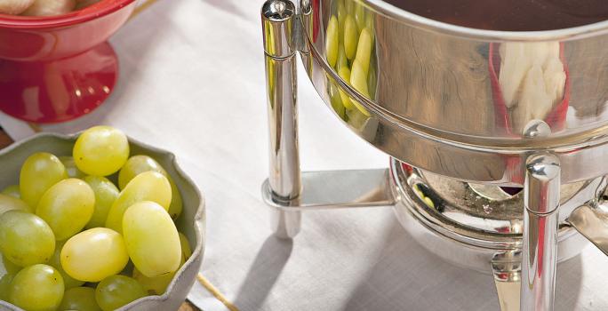 Fotografia em tons de verde e vermelho em uma bancada de madeira, uma toalhinha cinza, potinhos vermelhos com banana, uva, morango e abacaxi dentro deles. Ao centro, uma panela com a fondue de chocolate amargo dentro.