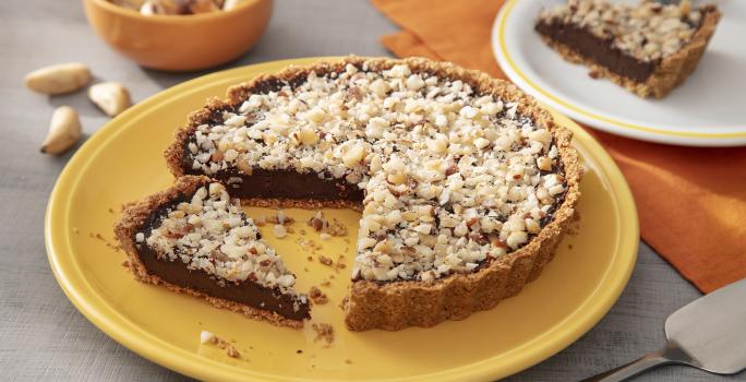 Fotografia em tons de amarelo em uma bancada de madeira de cor cinza. Ao centro, um prato amarelo contendo o cheesecake com uma espátula ao lado. Ao fundo, há um pano laranja com um pires branco em cima contendo uma fatia da sobremesa e algumas castanhas.