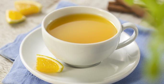 Fotografia em tons de azul, cinza e amarelo de uma bancada cinza com um paninho azul e sobre ele um prato branco redondo com uma xícara com chá de laranja. Ao fundo paus de canela e pedaços de laranja.