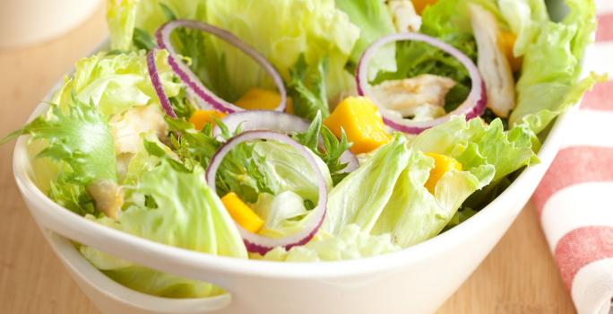 em uma mesa de madeira contém um recipiente branco que comporta a salada. Ao lado um pote com tempero e um pano listrado nas cores branco e rosa.