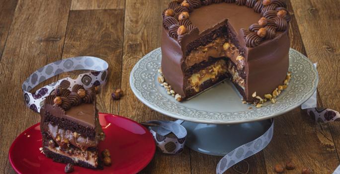 Foto da receita de bolo de chocolate com especiarias, sobre um prato cinza e sem uma fatia que está num prato vermelho do lado esquerdo. Tudo está em uma bancada de madeira decorada com uma fita de páscoa e pedaços de avelãs