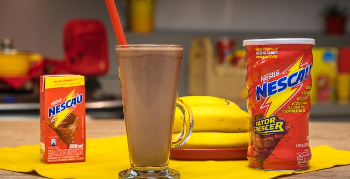 Fotografia em tons de vermelho em uma bancada de madeira vermelha com um copo de vidro ao centro e a vitamina de Nescau dentro dele. Ao fundo, um prato com três bananas.