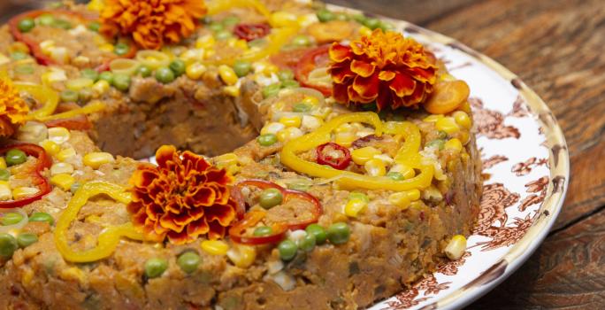 fotografia em tons de marrom e amarelo de uma bancada marrom vista de frente. Contém um prato redondo branco com um cuscuz, com pedaços de legumes e flores por cima para decorar.