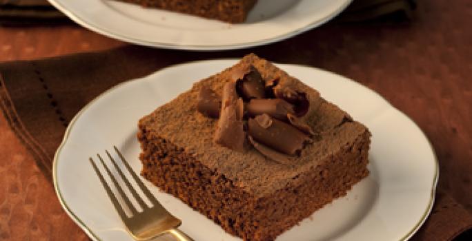 Fotografia em tons de marrom em uma mesa de madeira escura com dois pratos rasos brancos com dois pedaços do bolo de chocolate decorado com raspas de chocolate.
