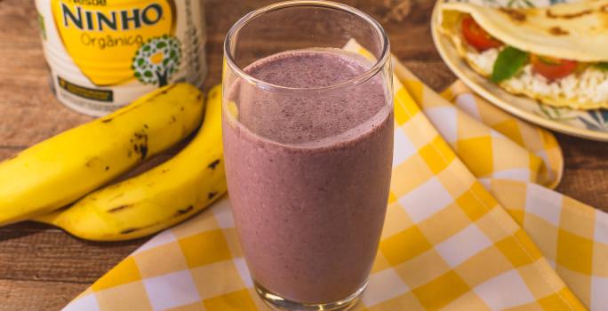 Fotografia em tons de amarelo de uma bancada com um pano xadrez amarelo e branco e um copo com a bebida de açaí. Ao fundo bananas, uma lata de NINHO e um prato com uma crepioca.