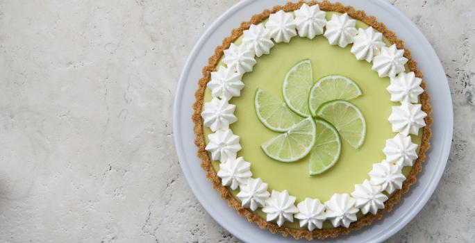 Foto de uma torta limão em cima de um prato branco e com fatias de limão decorando o centro da torta.