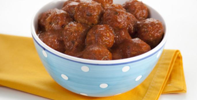 fotografia em tons de amarelo e branco, contém um pano amarelo e por cima um recipiente redondo azul com bolinhas brancas que contém bolinhos de carne com molho de tomate por cima