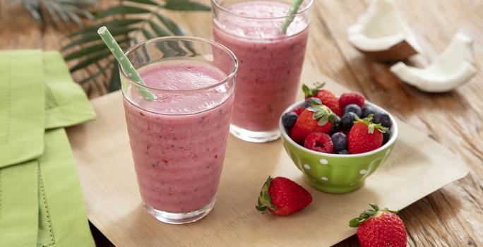 Fotografia em tons de vermelho, em uma bancada de madeira de cor marrom. Ao centro, uma bandeja de madeira com dois copos em cima contendo os smoothies. Ao lado, há um pano verde, uma tigela com morango e ao fundo algumas folhas e um coco partido ao meio.