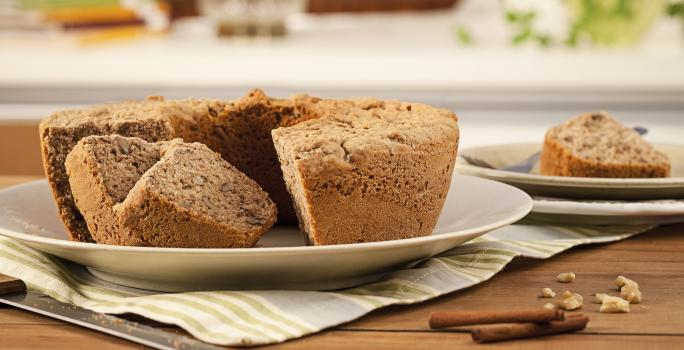 Fotografia em tons de marrom em uma mesa de madeira com um prato raso branco apoiado em um pano cinza listrado em bege e o bolo de maçã e nozes. Ao lado, um prato com um pedaço do bolo e paus de canela espalhadas pela mesa.