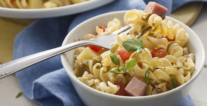 Fotografia em tons de azul em uma bancada de madeira com um pano azul ao fundo, um prato redondo fundo com a salada de macarrão tipo parafuso, com bacon e tomate.