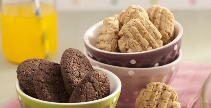 Fotografia em tons de rosa em uma bancada de madeira clara com potinhos verde, branco e marrom com bolinhas brancas em cima de um pano rosa e biscoitinhos de chocolate e aveia dentro dos potinhos.