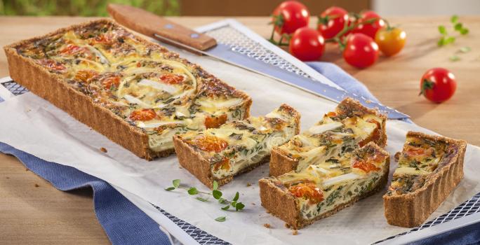 Fotografia em tons branco, dourado e vermelho, a direita e ao alto tomates-cereja e folhas de tomilho, ao centro travessa branca com quiche assada e fatiada, sobre papel manteiga e guardanapo azul em bancada de madeira.