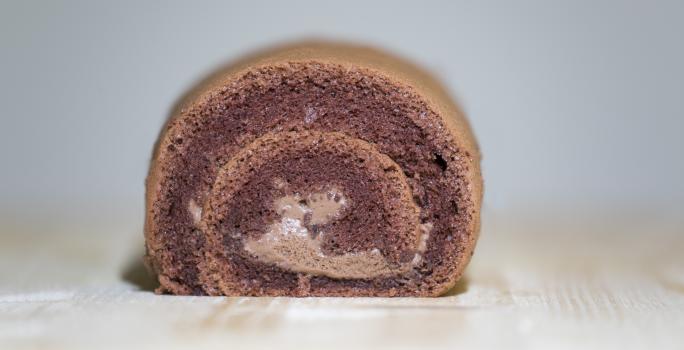 Fotografia de um bolo de rolo de chocolate, em tons de marrom, ao centro de uma mesa de madeira clara.