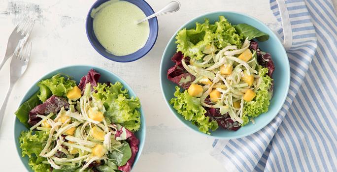 Fotografia em tons de branco, azul e verde de uma bancada branca vista de cima, contem dois potes azuis e redondos e ambos com saladas dentro. Ao lado um pano azul com branco, 2 garfos e um pote redondo azul com molho