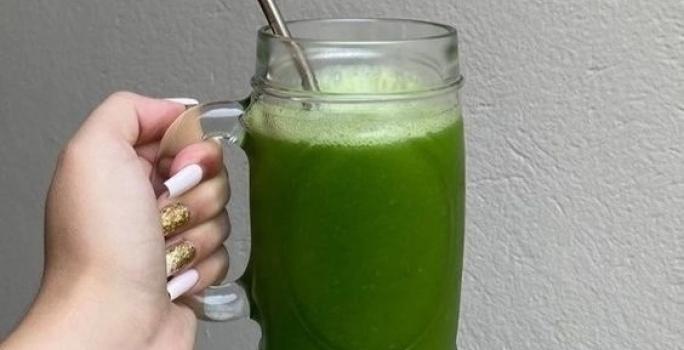 Fotografia de uma mão feminina segurando uma caneca de vidro grande com suco detox, de cor verde, com canudo de alumínio.