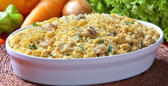 Fotografia em tons de amarelo em uma bancada de madeira de cor marrom. Ao centro, uma travessa branca contendo a farofa e ao fundo, alguns legumes e verduras espalhados.