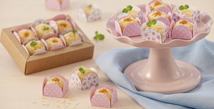 Fotografia em tons de rosa em uma bancada de madeira rosa com um pano azul, um suporte para bolo rosa com alguns brigadeiros brancos com geleia de maracujá em forminhas rosas. Ao fundo, uma caixinha bege com mais brigadeiros brancos.