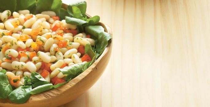 Fotografia de uma mesa de madeira com um bowl de madeira em cima com a salada dentro com folhas, feijão e cenoura.