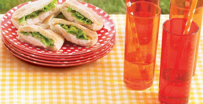 Fotografia em tons de vermelho e amarelo em uma mesa com uma toalha amarela xadrez, pratos vermelhos com bolinhas brancas e os sanduíches de presunto em cima deles. Mais à frente, copos compridos laranjas com suco e canudos de papel.