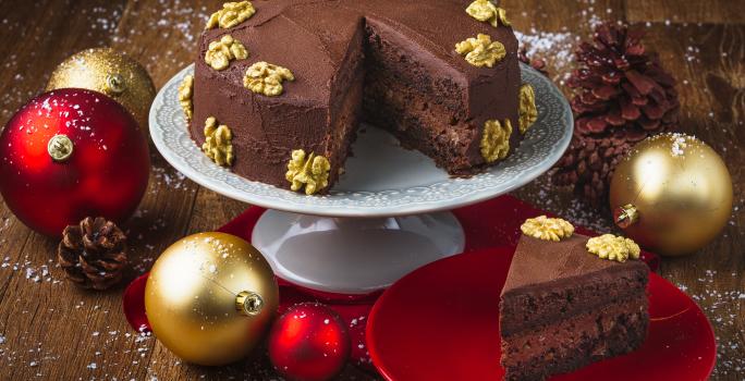 Foto da receita de Bolo Cremoso de Chocolate e Nozes, em um prato branco alto e uma fatia em um prato vermelho. Tudo está em uma bancada de madeira escura decorada com bolas de natal, pinhas e neve artificial