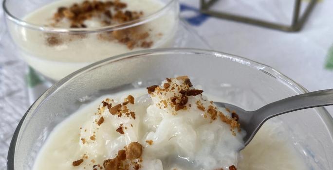 FotografiaFoto de um pote de vidro com a receita de Arroz Doce dentro, decorado com amendoins torrados. Há uma colher pegando um pouco do pote e ao fundo é possível ver a mesa decorada com uma toalha onde estão todos os itens da foto.