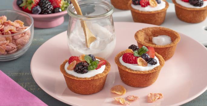 Fotografia em tons de rosa em uma bancada de madeira com um prato raso rosa ao centro com três cestinhas crocantes com frutas, iogurte e cereais em cima dele. Ao fundo, potinho rosa com frutas vermelhas.