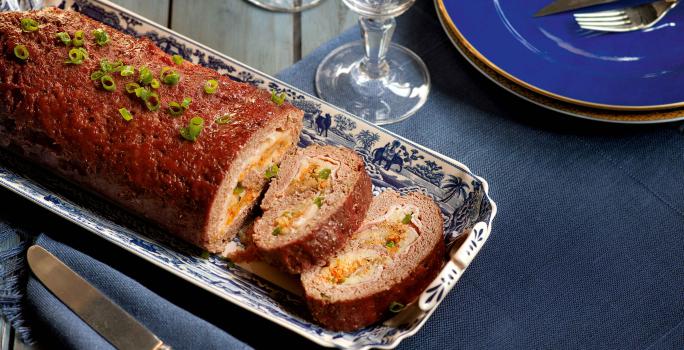 Fotografia em tons de azul escuro em uma mesa de madeira com uma travessa de prata ao centro com o bolo de carne recheado com vegetais Ao lado, um prato azul com garfo e faca apoiados nele e duas taças de vidro.