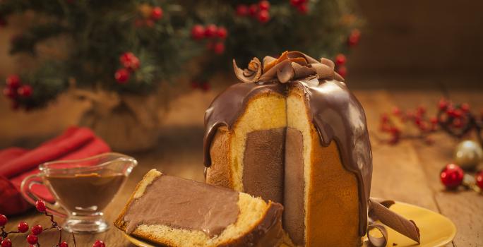 Foto frontal de um panettone com uma fatia cortada. Ele está sobre um prato amarelo e dentro há um recheio de sorvete em tom marrom escuro. Na bancada de madeira há elementos de natal e um pequeno recipiente com calda de chocolate