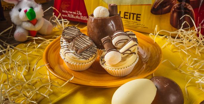 Foto aproximada de três miniovos de chocolate brancos, em um prato amarelo sobre uma bancada de madeira decorada com palha, um tecido amarelo, um pequeno coelho de pelúcia e uma barra de chocolate ao fundo