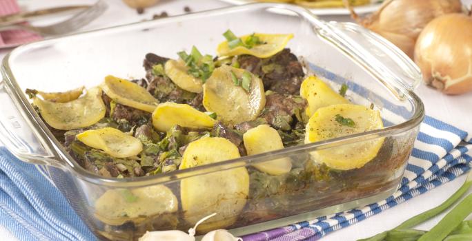 Fotografia de uma travessa de vidro quadrada com iscas de fígado e batatas, sobre um pano de mesa dobrado na cor azul com listras brancas. Ao fundo, cebolas e batatas inteiras, ao lado de um prato com legumes cortados.