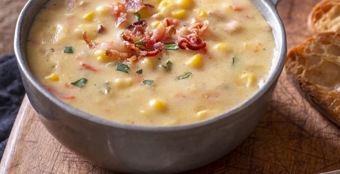 Foto aproximada de uma tigela cinza com a receita de sopa de milho em tom amarelo, sobre uma tábua de madeira ao lado de uma colher de metal.