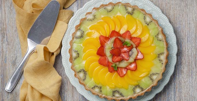 fotografia em tons de cinza, amarelo, verde e branco de uma bancada cinza vista de cima. Ao centro um prato redondo branco com uma torta com pedaços de frutas por cima para decorar, e ao lado um pano pardo cm uma espátula.
