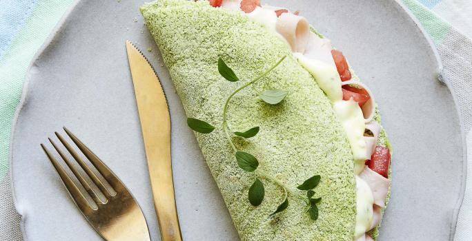 Fotografia em tons de verde em uma mesa de madeira com um pano listrado colorido em tons claro, um prato lilás redondo raso com uma tapioca verde de espinafre em cima dele.