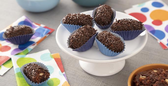 Foto da receita de Brigadeiro Croquete. Observa-se uma boleira pequena com 5 brigadeiros em formato de croquete em cima. A foto é decorada com guardanapos estampados de bolas coloridas e ao fundo observa-se dois potinhos: com brigadeiro e biscoito maria