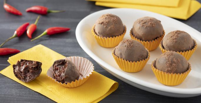 Fotografia em tons de amarelo de uma bancada cinza, sobre ela um prato oval com cinco trufas em forminhas amarelas, ao lado esquerdo um guardanapo amarelo com uma trufa cortada ao meio sobre ele. Ao fundo pimentas vermelhas.