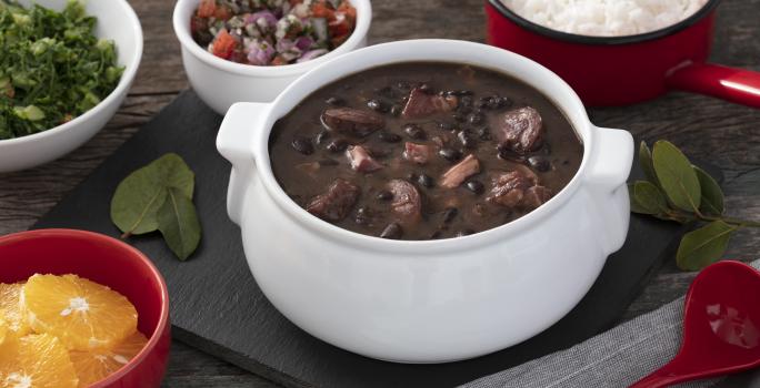 Fotografia em tons de vermelho em uma bancada de madeira escura, uma tábua preta com uma panela branca com a feijoada dentro dela. Ao lado, vários potinhos com laranja, couve, arroz e farofa.