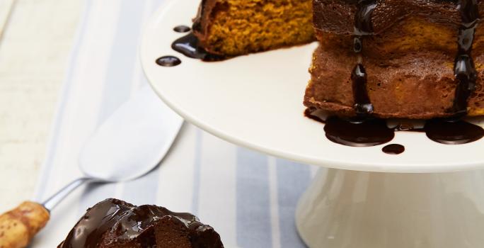 Fotografia em tons de marrom em uma bancada de madeira com um pano branco com listras azuis. Ao centro, um prato branco redondo raso com uma fatia do bolo de cenoura com chocolate. Ao fundo, um suporte branco para bolos com o bolo inteiro.