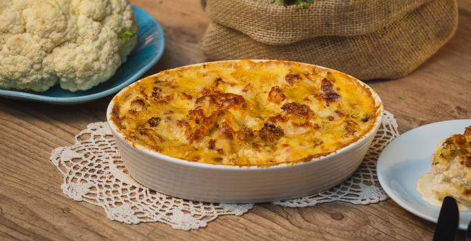 Fotografia em tons de amarelo em uma bancada de madeira clara, um paninho de renda e um recipiente branco oval com a couve-flor gratinada feita na AirFryer Mondial dentro dele. Ao lado, um prato com couve-flor crua, um outro prato com uma fatia.