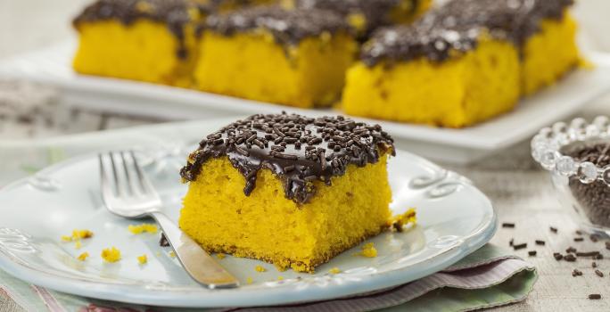 Foto de um pedaço de bolo de cenoura com cobertura de brigadeiro em um prato branco com um garfo do lado