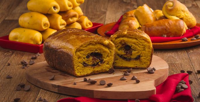 Foto de uma bancada de madeira. Sobre ela há uma tábua sobre um pano vermelho e nela está um pão em tom laranja aberto ao meio com recheio de chocolate. Ao fundo há mais pães menores e espalhadas pela bancada estão algumas gotas de chocolate